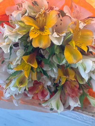 This media file features a close-up, overhead shot of a vibrant bouquet of alstroemeria flowers. The flowers are a mix of colors, including bright yellow, white, and soft orange or coral hues, with delicate dark speckles and streaks on some petals. Green foliage is interspersed throughout the bouquet, adding depth and contrast. The flowers appear fresh and are arranged tightly together, filling the frame. The background is blurred, with hints of orange wrapping material on the left and a light blue, possibly fabric or paper, surface on the right. The lighting suggests it might be daytime, possibly outdoors or near a window, as there are soft highlights on the petals. There is no text visible in the image. The overall impression is a colorful and cheerful display of flowers.