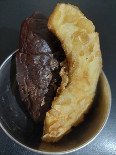 In a small, metallic bowl, two pieces of food are positioned side-by-side against a dark, matte background. The food item on the left is dark brown and has a rough, textured surface, suggesting it might be a piece of dark chocolate or a dense brownie. The food item on the right is golden yellow and appears to be fried, with a crispy, uneven texture, possibly a piece of fried banana or a fritter. The bowl itself is partially visible at the bottom and side of the frame, reflecting some light. The overall scene is a close-up shot focused on the food items. There is no indication of the city of Jelutung, Indonesia, in the image.