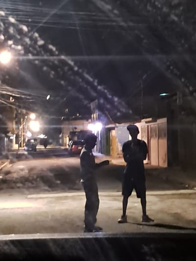 The image depicts a nighttime street scene in Cumana, Venezuela, likely a residential or commercial area given the presence of buildings and street lighting. Two individuals are visible standing in the middle of the street, silhouetted against the ambient light. The person on the left is facing towards the right, appearing to be speaking or gesturing with their hand. The person on the right is facing forward, with their arms crossed. Cars are present in the background, one appearing to be a red sedan. The overall atmosphere is dimly lit, with streetlights casting a glow on the wet or reflective street surface. The scene suggests a casual or perhaps impromptu gathering on the street at night.