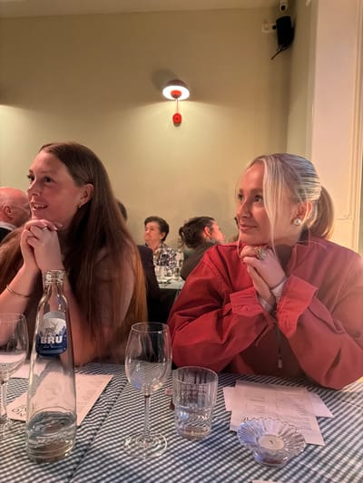 The image depicts an indoor dining scene in a restaurant or similar establishment located in Ixelles - Elsene, Belgium. Two young adult females are prominently seated at a table in the foreground, facing slightly right and looking towards an unseen area outside the frame. The woman on the left has long, straight, light brown hair and wears a light-colored top, with her hands clasped on the table. A small gold hoop earring is visible on her left ear and a thin gold-colored bracelet on her left wrist. The woman on the right has blonde hair pulled back in a ponytail and wears a rust-red oversized collared shirt over a white undergarment, also with her hands clasped on the table. She wears a large pearl earring on her right ear and a prominent gold-toned ring with a green stone on her right ring finger.

The table is covered with a green and white checkered tablecloth. Objects on the table include an empty glass bottle of "BRU" mineral water with a blue label depicting a stag, two empty wine glasses, an empty ribbed tumbler glass, and several white paper menus or documents. A small, fluted clear glass candle holder with an unlit tea light is also present.

In the blurred background, several other patrons are seated at tables. A bald male's head is partially visible behind the left woman, and an older female with dark hair is visible further back. The walls are a plain light beige. A red wall-mounted sconce with a visible pull cord emits light onto the wall, positioned above and between the two main subjects. In the upper right corner, a black ceiling-mounted security camera or speaker is visible. The overall lighting is soft and warm.