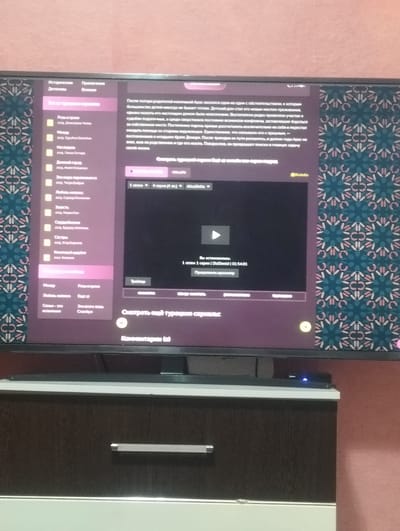 The image shows a television screen displaying a streaming service interface, likely a movie or TV show. The left side of the screen features a menu with categories like "Historical," "Detective," "Romance," and "Action." Under "Romance," there are sub-options such as "Rose and Thorns," "Between Us," "Between," and "Love of My Life." The right side of the screen presents a synopsis for a program, describing a character named Aras who is alone after the death of his parents and is looking for work. There's also a video player interface with playback controls, episode selection, and a "Play Trailer" button. The lower part of the screen shows "Comments (0)." The television is placed on top of a dark brown wooden cabinet with a silver handle. The background of the television screen has a patterned wallpaper in shades of teal and pink. There is no indication or visual information related to the city of Grinăuți, Moldova, in the image.
