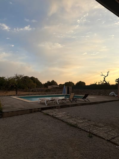 The image depicts a serene outdoor scene at dusk, likely in the vicinity of Santa Margalida, Spain. A rectangular swimming pool with clear blue water is the central focus, surrounded by a wooden deck and a gravel path. Several lounge chairs are positioned around the pool, some unoccupied and others with a white umbrella providing shade. In the background, a stone wall marks the property boundary, beyond which trees and vegetation are visible under a sky painted with the warm hues of sunset. A stark, bare tree silhouette stands against the horizon, adding a touch of natural drama to the landscape. The overall impression is one of tranquility and relaxation, with the natural light of the setting sun casting a soft glow over the scene.