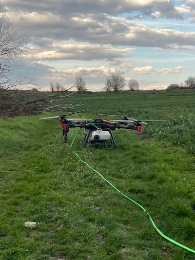 An agricultural multi-rotor drone is positioned centrally on a field of green grass and low-lying vegetation in Ruska Bela, Bulgaria. The drone is predominantly black with red accents on its arms and features a white liquid tank mounted underneath its main body. Four visible propeller blades are black. A bright green hose, with sections striped in yellow, is connected to the drone and extends diagonally across the grass towards the lower-right corner of the frame. To the left of the drone, a pile of cut dry tree branches is visible on the ground. In the background, a line of leafless deciduous trees stands against the horizon. The sky is partly cloudy, displaying a mix of grey and white stratocumulus clouds with patches of blue. A small, light-colored object is partially visible within the grass in the lower-left foreground. No individuals are present in the scene.