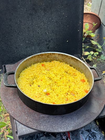 A wide shot from above shows a pot of yellow rice with chunks of red bell pepper and other ingredients cooking over an outdoor fire pit. The rice fills a large, silver-colored pot with two black handles. The pot is situated on a dark, circular metal structure, which is part of a rustic outdoor cooking setup. Coals and embers are visible to the right of the pot, indicating the fire is actively burning. The background includes a dark, textured surface on the left, possibly a table or stovetop, and some greenery and a terra cotta pot to the upper left, suggesting an outdoor setting. The scene appears to be taking place outdoors during the daytime, with natural light illuminating the food. The focus is on the prepared rice, suggesting a meal is being cooked or has just been finished.
