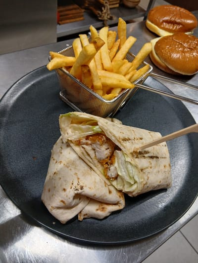 The image displays a meal composed of a chicken wrap and french fries, presented on a dark grey, textured circular plate. The chicken wrap is diagonally sliced, revealing its interior of crispy, breaded chicken pieces, shredded green lettuce, and a creamy white sauce. The tortilla shell exhibits distinct char marks. A light-colored wooden skewer is inserted into the top half of the wrap. Adjacent to the wrap, a small, silver-colored wire mesh basket, equipped with two handles, contains a portion of golden-brown french fries. In the blurred background, resting on a reflective, scratched metallic surface, are two brioche-style buns, one appearing to contain a light-colored filling or cheese. Further in the background, indistinct brown objects, possibly wood or rope, are visible. The overall scene is set on a stainless steel counter surface, with a white tiled floor partially visible in the bottom right, indicative of a commercial kitchen or food preparation area in the city of Sotira, Cyprus.