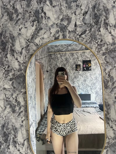 In the city of Blizhnii Khutor, Moldova, a young woman stands in front of a large, oval mirror with a gold frame. The mirror reflects a bedroom with a bed covered in a patterned duvet and a pillow that appears to have the text "CHRISTIAN DIOR" on it. The walls are covered in a grey and white marble-patterned wallpaper. The woman is holding a smartphone in her right hand, taking a selfie. She is wearing a black crop top and leopard-print shorts. Her long, dark hair falls over her shoulders. On the wall behind her, visible in the mirror, are two small framed artworks. One contains handprints of various colors, and the other features handprints and footprints. A tan-colored cloth hangs to the left of the mirror.