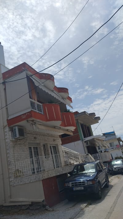 In Νέα Μηχανιώνα, Greece, a street scene unfolds under a cloudy sky. On the left, a multi-story building with distinctive orange balconies and white awnings exhibits signs of wear. An air conditioning unit is visible on the first floor. Adjacent to this building, a dark blue pickup truck with the license plate NHY 4997 is parked on the asphalt road, partially obscuring the lower level of the building. Further down the street, a dark-colored car is also parked. The road appears to be a typical urban street with some debris and vegetation on the side. The buildings in the background suggest a residential or commercial area.