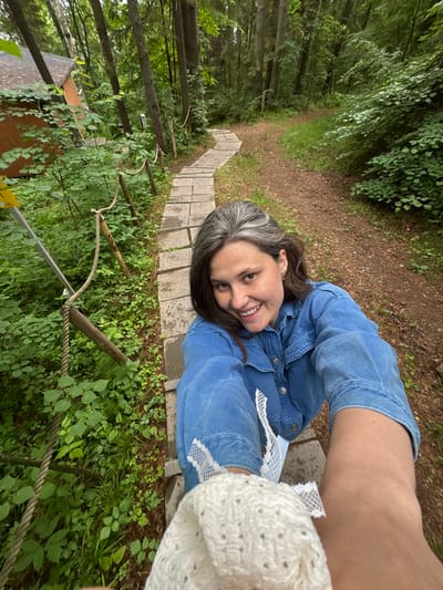 A woman with graying hair, wearing a blue denim shirt and holding a white knitted item, smiles at the camera while taking a selfie. She is positioned on a path made of stone slabs in a forest. To her left, a wooden cabin with an orange roof is visible amongst dense green foliage and trees. A rope railing runs alongside the path, supported by wooden posts, further into the wooded area. The ground beside the path is a dirt trail covered with fallen leaves and grass. The overall environment suggests a natural, possibly recreational or residential setting within a forest.