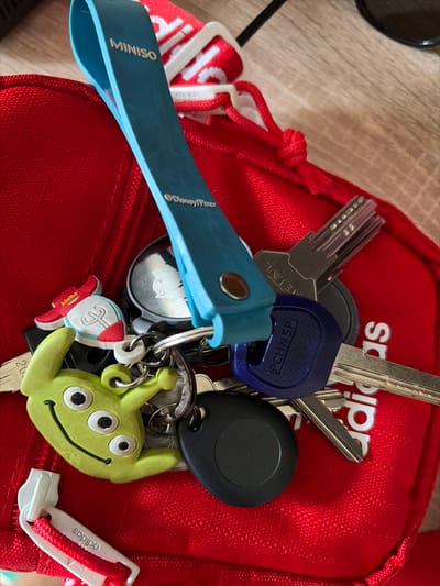 A close-up, eye-level shot shows a red fabric bag with a zipper pull attached to a keychain. The keychain consists of a blue Miniso branded strap, several keys, and three keychains: a three-eyed green alien from Toy Story, a red and white rocket-shaped keychain, and a circular black tag. The bag is on a wooden surface. The image does not provide any information about the city of Svetlia, Bulgaria.