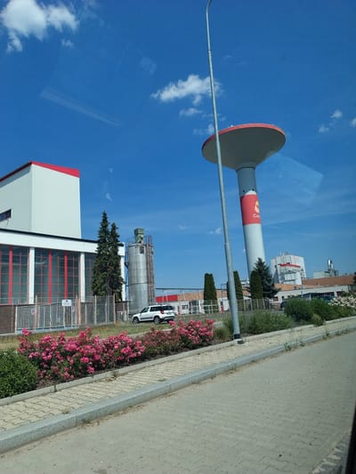 The image shows an industrial area with a tall, cylindrical water tower with a red rim and a red band with the word "Culigo" printed in white. A white SUV is parked on the right side of the road, and a building with a white facade and red trim is visible on the left. There are also several trees and a row of pink rose bushes in the foreground. The sky is blue with some white clouds. The scene appears to be located in Horažďovice, Czechia.