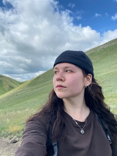 A young woman with long, dark, wavy hair, wearing a black beanie, a brown long-sleeved shirt, and a necklace, is pictured in a selfie. She has a nose ring and distinctive earrings. She is outdoors, with rolling green hills and a cloudy sky in the background. The image appears to have been taken in the city of Andreevka, Russia.