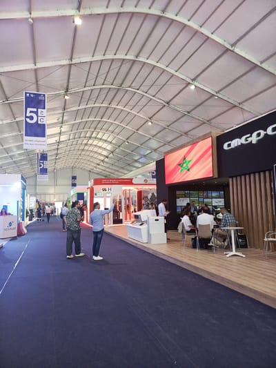 The image captures an indoor scene from a bustling trade show or exhibition held within a large white tent structure. The tent features a high, arched ceiling with visible metal supports and evenly spaced artificial lighting fixtures. A dark blue carpet covers the central aisle, guiding visitors through the event.
Numerous people are visible, some walking along the aisle, others gathered at booths. In the mid-ground, two men stand facing away from the camera, observing the exhibits. One wears a patterned shirt, and the other, in a light blue shirt, gestures towards a booth. On the right side, a group of approximately five to six individuals are seated around small white tables at a large, prominent exhibition stand, engaged in conversation, suggesting business discussions or networking.
This main booth on the right features a large LED screen prominently displaying the flag of Morocco, immediately identifying the location as Morocco. Above this screen, the company name "CMGP-C" is displayed in large white letters on a dark background. Further back along the right, another booth features the text "Leader en fabrication et commercialisation des tuyaux en Plastique" alongside "H" and "HOR Iud," indicating a focus on plastic pipe manufacturing. On the left side of the aisle, a booth with "ROTA-PLAST" is visible, and further down, elements of "GEMAPL" are discernable.
Multiple blue banners are suspended from the tent ceiling. These banners feature "5 ج / C" and the Arabic text "المستلزمات الفلاحية" (Agricultural Supplies), along with its French translation "Pôle Agro-fournitures" (Agro-supply hub), suggesting the event is focused on agricultural supplies or related industries. The overall atmosphere is lively and professional, characteristic of a daytime trade event in Al Machouar – Stinia, Morocco.