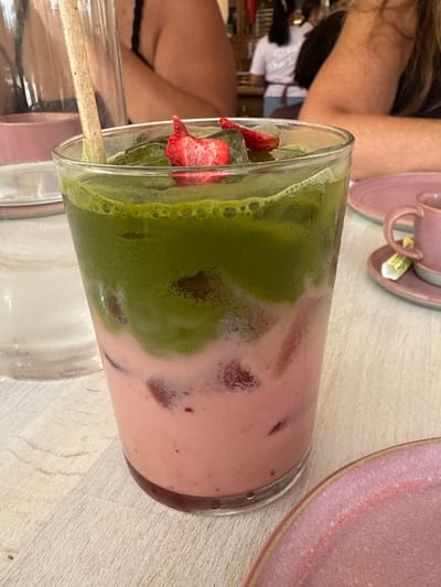 The scene is an indoor dining establishment located in North Miami, United States. The primary subject is a clear glass containing a two-layered beverage, positioned on a light-colored table surface. The lower liquid layer is opaque pink with embedded red solids, consistent with fragmented strawberries. The upper layer is opaque green, consistent with matcha, containing ice cubes and two irregular, rehydrated red strawberry pieces floating on the surface. A light-colored wooden straw is inserted into the drink. To the right, a pink ceramic plate and a matching pink cup on a saucer are visible, with a small, wrapped white packet resting on the saucer. To the left, a portion of a clear glass containing water is present. The background, out of focus, reveals two individuals partially seated at tables; one on the far left wears a dark-colored top. A third individual, likely an employee, stands further in the background near a service counter, wearing a light-colored top and a dark apron. The environment is consistent with a casual cafe or restaurant setting.