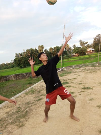 A young male, barefoot and wearing a black t-shirt and red shorts with the number 42 on them, is captured in mid-action, playing volleyball. He is positioned on a dirt court, with a volleyball net set up behind him. The background reveals a lush, green landscape of rice fields, typical of the rural scenery found in Turumame, Indonesia. Several palm trees and a few simple wooden houses are visible further in the distance, suggesting a tranquil, countryside setting. The sky is partly cloudy, indicating an outdoor activity taking place during the day.