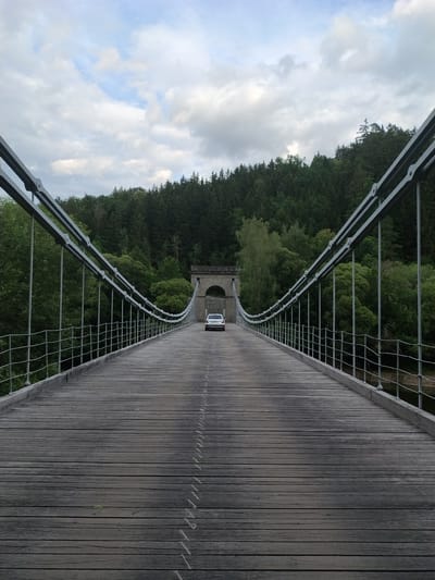 A white car is driving across a wooden suspension bridge. The bridge has metal railings and suspension cables. The bridge is surrounded by trees, and there is a cloudy sky overhead. The location is the Stádlec Bridge in Stádlec, Czechia.