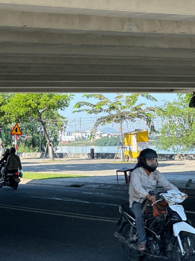 The image depicts a street scene in Da Nang, Vietnam, under an elevated concrete structure, likely a bridge or overpass. In the foreground, a person wearing a light-colored long-sleeved shirt, blue jeans, and a black helmet is riding a white and black motorcycle. The motorcycle has an orange accent visible beneath the rider. To the left, another motorcycle with two people wearing darker clothing is also on the road.

Beyond the road, a paved walkway lines a body of water, possibly a river or bay. Green trees and foliage are abundant on the other side of the walkway. A yellow kiosk with the words "KEM DUA MALAI" written on it, along with posters, is situated near the water's edge. A red traffic sign with a yellow triangle and a black pictogram of a pedestrian and a cyclist is visible on the left side of the scene. In the background, buildings and structures are visible across the water, and overhead, power lines and decorative lights hang from poles. The lighting suggests it is daytime, with sunlight casting shadows on the road and ground.