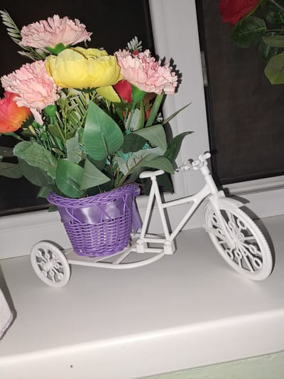 This is a close-up, eye-level, indoor shot of a decorative white plastic bicycle with a woven purple basket filled with artificial flowers and greenery, placed on a white ledge against a dark background. The bicycle is a tricycle with a front wheel and two smaller rear wheels. The basket is decorated with a purple ribbon. The flowers include pink, yellow, and orange blooms, along with green leaves. The ledge appears to be a windowsill. The overall scene is a still life arrangement. The location is specified as Ынтымақ ауылдық округі, Kazakhstan.