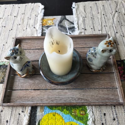 The image shows a rustic wooden tray on top of a cream-colored woven placemat with black stitch details. Inside the tray, there are two ceramic cat figurines and a thick, cream-colored pillar candle in a dark blue ceramic dish. The cat figurine on the left is facing forward and decorated with blue and white stripes and floral patterns. The cat figurine on the right is facing to the right and decorated with blue floral patterns and brown and orange flowers on its head. The candle is lit and has melted wax around its edges. The placemat has tasseled edges and is placed on a dark surface, possibly a table. In the background, part of a jigsaw puzzle with a colorful nature scene is visible. The scene is indoors, and the lighting suggests it could be daytime with natural light. The image is taken from a slightly overhead perspective. The location of this scene is unknown and cannot be determined from the image.