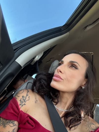 A woman with dark hair and fair skin is in a car. She is wearing a seatbelt and a red top. She has tattoos on her shoulders and chest and is wearing a diamond necklace and earrings. Her sunglasses are placed in her hair. The location in the city of Kazachevo, Bulgaria, cannot be verified.