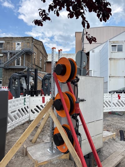 This media file shows a construction site in Mannheim, Germany. The scene is outdoors with a cloudy sky overhead. The perspective is from a slightly elevated angle, looking down and to the right.
Dominating the foreground are two large orange cable reels mounted on a red metal frame. The cables are black and appear to be heavy-duty electrical cords. These reels are suspended above a grey metal structure with multiple horizontal bars. Below this structure, two forklifts are visible. They are dark grey and red, with visible "RJHS" branding on their cabs.
In the background, there are several modular building units, likely temporary site offices or storage containers. These units are white and grey with corrugated metal siding and windows. A brick building with an external metal fire escape staircase is also visible to the left. A dark-leaved tree branch extends into the frame from the upper left, silhouetted against the sky.
The ground is a mix of gravel and concrete, with some wooden planks and white plastic barriers also present, suggesting ongoing construction work. The overall impression is one of active construction and utility work. There are no people visible in the image. The time of day appears to be daytime, given the brightness of the sky, though the clouds suggest it might not be a perfectly sunny day.
