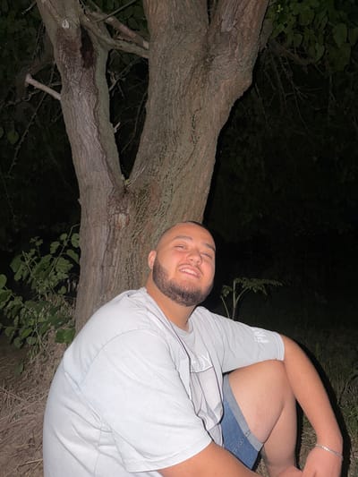 In the city of Debelets, Bulgaria, at night, a young man with a shaved head and a beard is sitting near a tree. He is wearing a light-colored t-shirt and denim shorts. The man is smiling at the camera, with his left arm resting on his knee. The background is dark, with the tree trunk and branches clearly visible. There is some foliage to the left of the man, and the ground is covered in dry grass and twigs. The lighting suggests a flash was used to illuminate the scene.