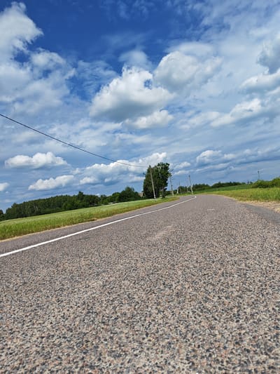 The image depicts a rural landscape under a dynamic sky with dramatic clouds. A paved road with a white line marking its edge curves into the distance. On either side of the road are fields of green grass and dense trees. Several utility poles with wires are visible along the roadside, and a single tree stands prominently on the left. The scene appears to be located in a rural area, possibly the Ленінскі сельскі Савет in Belarus, given the context. There are no people or animals visible in the image. The overall impression is one of a clear, bright day with a sense of open space and natural beauty.