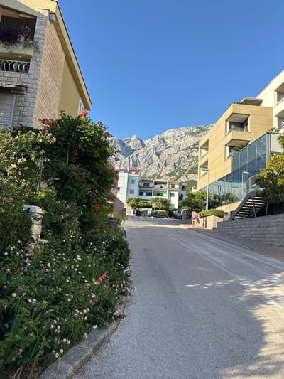 The image depicts a street scene in Makarska, Croatia, with a paved road leading uphill towards a range of rocky mountains under a clear blue sky. On the left, a building with a yellow facade is partially visible, adorned with abundant green foliage and red and pink flowers. On the right, a modern building with a beige facade features extensive glass panels and balconies. Two individuals, one appearing to be a woman in a light-colored dress and the other a person in darker attire, are walking uphill on the sidewalk. The overall environment suggests a warm, sunny day.