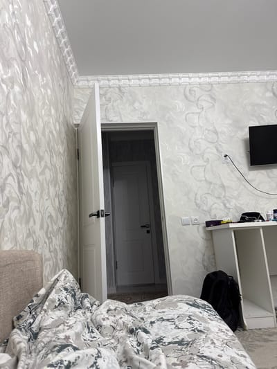 The image shows an indoor scene, likely a bedroom or living space, featuring a white door ajar, revealing another doorway. The walls are adorned with ornate, light-colored wallpaper with a swirling pattern. A white ceiling molding runs along the top of the walls. In the foreground, a section of a bed with patterned duvet is visible, along with a portion of a sofa on the left. To the right, a white desk is present, with a black backpack leaning against it. A black television is mounted on the wall above the desk, and electrical outlets and a black cord are also visible. The overall impression is of a tidy but lived-in domestic environment. This location is in the city of Zhanaozen, Kazakhstan.