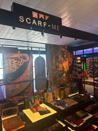 This image presents an interior view of a retail establishment named "SCARF • ME," situated in an indoor commercial environment, likely an airport or shopping center in Guarulhos, Brazil. This location is suggested by the visible "Kibon" brand freezer in an adjacent store.

A prominent black sign, bearing the white capital letters "SCARF • ME" and three illuminated orange flag-like symbols, is suspended from the ceiling in the upper portion of the frame. The store specializes in scarves, which are extensively displayed.

On the left, large scarves featuring a variety of designs, including a prominent golden jellyfish against a dark, starry background and vibrant botanical patterns, are hung against a large window. This window allows natural light to enter and reflects exterior buildings. To the right of these hung scarves, a tall, oval mirror with a gold frame stands upright.

The foreground and midground consist of black display cases and tables, some featuring illuminated white platforms, showcasing a wide array of folded scarves. These scarves exhibit diverse patterns, including intricate floral, geometric, abstract, and animal print designs, in various color palettes. Several woven straw bags or purses are also displayed, along with a "SCARF • ME" branded shopping bag featuring a botanical design. A dark mannequin bust is partially visible among the displays on the right side.

In the background, through glass partitions, an adjacent retail store is visible. This store features shelves stocked with packaged snacks, bottled beverages, and a distinctive red and white "Kibon" brand freezer. The ceiling throughout the depicted area is constructed from white square acoustic tiles, with various recessed and surface-mounted lighting fixtures providing illumination. No individuals are present in the scene. The overall lighting is bright and consistent.