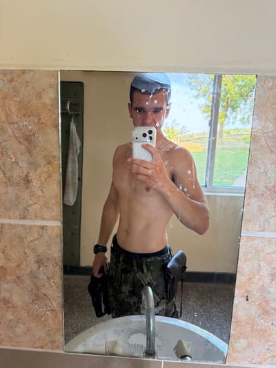 The image shows a man taking a mirror selfie in a bathroom. He is shirtless and wearing camouflage patterned shorts and a black watch. He has a towel or washcloth on his head. He is holding a smartphone in his right hand, which is pointed towards the mirror. The mirror is dirty and has water droplets on it. In the reflection, we can see a window with trees and sky outside, suggesting it is daytime. The bathroom appears to have tiled walls and a sink with a faucet. There is a towel hanging on a rack below the mirror. The man's expression is neutral. The location is identified as Momin Sbor, Bulgaria.
