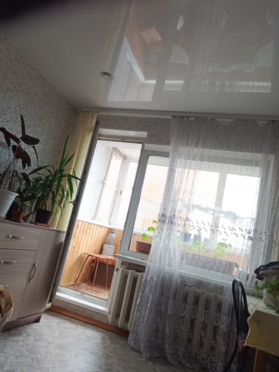 The image shows an interior view of a room with a large window and a sliding glass door leading to a balcony. The room appears to be a living area or bedroom, furnished with a dresser, a radiator, and a table with a chair. Several potted plants are visible on the windowsill and on the balcony. The decor is simple, with patterned wallpaper and a tiled floor. The lighting suggests it is daytime, with natural light streaming in through the windows. The room is located in Gusinoozyorsk, Russia. There are no people visible in the image, and no actions or interactions are taking place.
