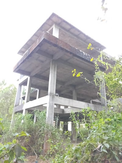 The image depicts the unfinished concrete framework of a multi-story building under construction in Marna, India. The structure is exposed, showing beams, columns, and floors without walls. Lush green vegetation surrounds the building, partially obscuring its view. The sky is bright, suggesting daylight conditions. No people or additional objects are discernible.