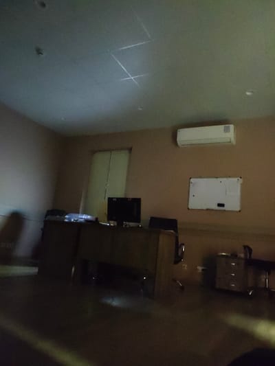 The image depicts an indoor office scene with subdued lighting. A prominent wooden desk with a computer monitor and a printer is visible in the foreground, along with office chairs. A whiteboard hangs on the wall above a white air conditioning unit. To the left, a closed door with blinds is partially visible. The room's walls are a neutral brown color, and the ceiling has a grid pattern with some light fixtures. The overall impression is of a functional, albeit dimly lit, office space. The location is specified as Sartichala, Georgia. There are no individuals or any actions or interactions taking place.