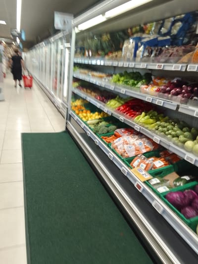 The image depicts the interior of a brightly lit supermarket aisle, with a focus on a display of fresh produce. In the foreground, a dark green mat runs along the bottom of the frame, adjacent to a cream-colored tiled floor. To the right, a refrigerated display case showcases an array of colorful vegetables and fruits on multiple shelves. Visible items include red and yellow bell peppers, green peppers, broccoli, cucumbers, onions, carrots, and red onions. Some produce is packaged in plastic, while others are loose. A price tag or label is visible on some of the produce containers.

Further down the aisle, a woman wearing a black dress is walking away from the camera, pushing a red shopping cart. She is partially obscured by the angle and blur of the image. The background shows more supermarket shelving and the reflective surfaces of refrigerated display units. The lighting is consistent with an indoor retail environment, suggesting it is daytime. There are no discernible external weather cues. The overall atmosphere is one of a typical grocery shopping experience. No visible text is clearly legible.