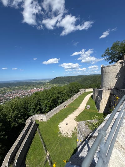 The image shows a view from a high vantage point, likely a castle or fortified structure, overlooking a valley with a town. The foreground features a stone wall and a metal railing, indicating the viewpoint is from within or adjacent to a historical building. A pathway winds its way down from the viewpoint, bordered by grassy slopes and stone walls. In the distance, a town with red-tiled roofs is nestled amongst rolling green hills and trees. The sky is blue with scattered white clouds. A small, reddish-brown dog is visible on the pathway. The location is identified as Neuffen, Germany, and based on the architecture and the prominent hill, it is likely the Hohenneuffen Castle.