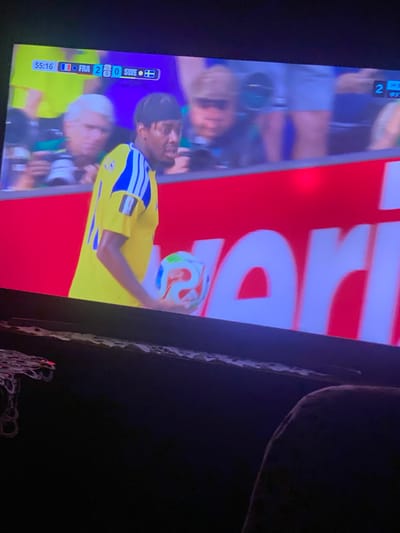 The image captures a sports match on a television screen. A football player in a yellow jersey with blue stripes is holding a multi-colored football. A scoreboard at the top of the screen shows "55:16" for the time, "FRA 2" for France, and "SWE 0" for Sweden, indicating the score and time of the game. The background shows blurred spectators and a red banner with white lettering. The television is placed on a dark surface, possibly a piece of furniture, and a person's leg is visible in the lower right corner, suggesting the image was taken from the perspective of an audience member. The scene is indoors, with the television screen being the primary focus. There is no indication of the location within the city of Zastavna, Ukraine.