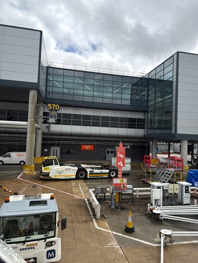 The image depicts an airport apron at Gatwick Airport, located near Crawley, West Sussex, United Kingdom.
The scene showcases an active airside area adjacent to a multi-story airport terminal building under a predominantly overcast sky. The modern terminal structure features large sections of dark-framed glass panels and alternating light grey and white facade panels. An enclosed glass skybridge connects two sections of the building at an upper level. Below this, a prominent yellow digital display reads "570," likely indicating a gate number. On a lower level of the terminal, an "easyJet stores" sign with orange branding is visible above an entrance. Large concrete pillars support the terminal structure, with one pillar in the foreground exhibiting yellow base markings.
On the concrete tarmac, various airport vehicles and ground support equipment are present. A white Ford Transit-style van, marked with "easyJet" and "engineering" logos, is parked on the left side of the frame. In the middle-ground, a white and yellow aircraft pushback tractor, identified as a "PHOENIX E" "AST-2" model with unit number "937" and a green bird logo, is positioned diagonally. In the foreground, the front section of a white and grey aircraft pushback tractor, branded "TREPEL" with a blue "M" logo and unit number "609832", is visible from an elevated perspective.
Ground support equipment includes several sets of red and silver mobile passenger stairs or work platforms staged on the right. Various cargo containers and carts are scattered across the tarmac. Red and white instructional signs, indicating "Emergency Stop Buttons," "Fuel Hydrant," and "Aircraft," are mounted on a stand near a cargo container. Multiple ground power units, one labeled "ATA POWER," along with associated cabling and hoses, are present. A yellow and black striped traffic cone rests on a stack of black rings on the tarmac. The concrete surface exhibits painted white and red demarcation lines defining operational zones. The sky is covered by grey, broken clouds, with minor patches of brighter sky. No people are visible within the frame.
