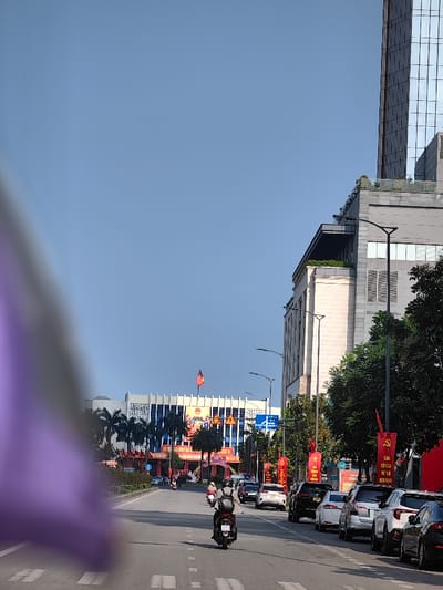 The image captures a street scene in Thuận Hóa Ward, Vietnam, under a clear blue sky. A blurry purple and grey object partially obscures the lower-left foreground. A paved road with dashed white lane markings extends into the distance. A single individual wearing a helmet and a backpack operates a dark-colored scooter in the left lane, moving away from the viewer. Multiple cars, including white, black, and grey sedans and SUVs, are parked parallel to the curb on the right side of the road. Additional vehicles are observed in motion further down the street.
Along the right side of the street, a multi-story building with a light-colored facade and a rooftop garden is visible, partially adjacent to a taller, modern building featuring a glass facade. Black, curved streetlights are positioned along the roadside. Numerous red banners with yellow Vietnamese text are vertically displayed on poles lining the right sidewalk. Green trees are interspersed along the street.
In the background, a large building with a prominent white or light-colored facade, characterized by vertical architectural elements and blue windows, stands centrally. A red and yellow Vietnamese national flag flies from its rooftop. An elaborate red and yellow decorative archway or structure is situated directly in front of this building.