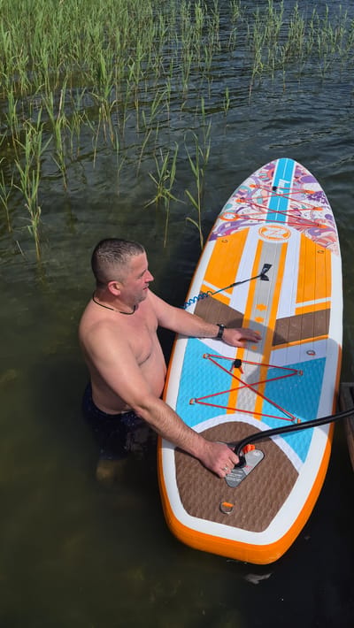 A shirtless man is standing in murky green water next to a colorful stand-up paddleboard. He is wearing dark patterned swim trunks. The man is reaching down with his right hand, touching the paddleboard. His left hand is also on the paddleboard. The paddleboard is white with orange, blue, and purple decorative patterns. A black hose is attached to the paddleboard. In the background, tall green reeds grow out of the water. The scene appears to be a natural body of water, possibly a lake or river, near vegetation. The man is facing to his left, looking away from the camera. The lighting suggests it is daytime.