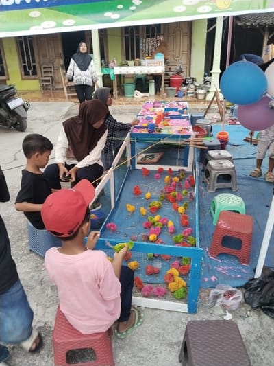 In Plosorejo, Indonesia, an outdoor fair is in progress, characterized by a lively scene of children and adults gathered around game stalls. A prominent stall features a blue wooden enclosure filled with vibrantly colored baby chicks – pink, yellow, green, and red – attracting the attention of young participants. Children are actively engaged in a game, attempting to catch the chicks with what appear to be small fishing rods. One boy, wearing a red cap, is holding a green chick, his attention focused on the game. Another boy, in a black t-shirt, sits on a stool, observing the activity. Adults, including women wearing hijabs, are supervising and managing the game. The area is laid out on a blue tarp, with various stools of different colors and materials placed around the stalls. In the background, a motorbike is parked, and a glimpse of buildings suggests a typical Indonesian street scene. Balloons, one blue and one purple, add to the festive atmosphere. The environment appears somewhat dusty, indicative of an outdoor event.