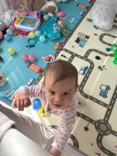A baby in a pink and white floral patterned long-sleeved shirt is near the edge of a crib, on top of a play mat with a road design. The play mat has two distinct areas: one with a city street pattern in beige and another with a blue base color. The blue section is scattered with various toys, including a white television toy, a plush dog, and colorful plastic balls. It is not possible to infer the location in the city of Veghel, Netherlands, from the image.