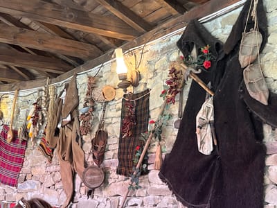 The image captures a wall adorned with various rustic items, suggestive of a historical or ethnographic display, possibly in Zheravna, Bulgaria. The wall is constructed of rough, light-colored stones. Above the wall, a wooden beamed ceiling is visible. Hanging from the wall are several objects. On the far right, a large, dark, shaggy cloak with a hood is displayed, from which a pair of small, light-colored leather shoes are suspended by twine. To the left of the cloak, there are red chili peppers strung together and a wooden object resembling a fork or rake. Further to the left, a dark plaid fabric hangs, and beneath it, a circular wooden object, possibly a drum, is suspended. To the left of that, a brown garment, possibly a vest or tunic, is displayed. Several bunches of dried garlic or onions are also visible. To the far left, a red and black plaid blanket or cloth is hung. A rustic oil lamp is mounted on the wall between the plaid fabric and the chili peppers. A wooden pole with green leaves and red roses entwined around it leans against the wall, passing behind the plaid fabric. The overall impression is that of a preserved historical setting or a themed exhibition.