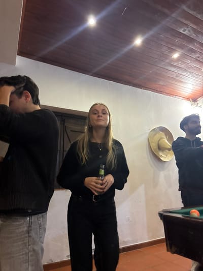 Three people stand in a room with a wood ceiling and white walls. The woman in the center looks at the camera, holding a drink. To her right is a pool table. Above her, a straw hat hangs on the wall. To her left, a man in a black sweater looks down. At the top of the frame, another person looks directly down. The room is illuminated by several bright lights in the ceiling.