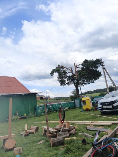 The image depicts a rural scene in the village of Islambaevo, Russia. The foreground is filled with various pieces of lumber, logs, and a partially assembled wooden structure, suggesting ongoing construction or woodworking. A bicycle lies on its side, with its front wheel propped up by lumber. Further back, a green building with a red roof is visible, with some potted plants near it. A bright yellow, small playhouse or shed stands in the yard. A white car, identified by its license plate "0716AX 702", is parked on the right side of the scene. In the background, a tall, distinctive pine tree with a gnarled trunk and branches dominates the landscape, with power lines and utility poles nearby. A green wooden fence runs across the middle ground, separating different areas of the property. The sky is a mix of blue and white clouds. The overall impression is one of a work in progress in a rustic, possibly summer setting.