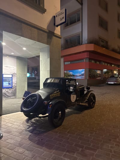 A dark-colored vintage roadster, emblazoned with "BULLOCK," a white racing number "1," and an orange "7," is parked on a brick-paved surface in Sankt Moritz, Switzerland. A spare tire is mounted vertically on the rear of the vehicle, which displays a Liechtenstein license plate "FL 2 287." The car's interior shows visible navigation equipment.

To the left, the entrance of a building, faced with light-colored stone panels, houses an ATM with a blue interface and an interior wall mural depicting a snowy mountain landscape. Above this entrance, a sign for "Graubündner Kantonalbank" is affixed.

To the right of the vintage car, a building features a lower section with red-paneled walls and windows displaying large posters of scenic mountain and lake vistas. Above this, the building's upper facade is light-colored stucco with rectangular windows.

Further in the background, to the right, a silver classic Porsche 911, bearing a Zurich canton license plate "ZH 901 199," is parked. Through a distant archway, illuminated decorative elements resembling blue Christmas trees and a sign for "GLATTFELDER" are discernible, along with a "10 km/h" speed limit sign. The scene is illuminated by artificial light, indicating nighttime. No individuals are present.