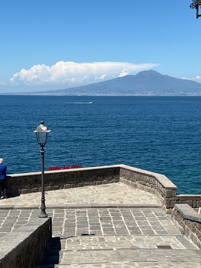 The image depicts a scenic view of the Bay of Naples from Sorrento, Italy, with Mount Vesuvius prominently in the background. In the foreground, a stone-paved pathway with a low stone wall overlooks the deep blue sea. A vintage-style lamppost stands on the right side of the pathway. Red lettering on the wall reads "Veuve clicquot dal mare". A person wearing a blue shirt stands at the left edge of the image, looking out at the sea. A distant boat is visible on the water, and a large cloud formation hovers above Mount Vesuvius. The sky is a clear, bright blue.