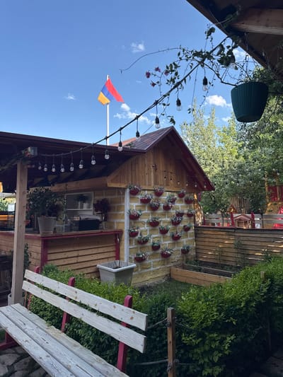 The image captures an outdoor seating area at a cafe or restaurant, likely in the city of Tsakhkadzor, Armenia, given the prominent Armenian flag flying on a flagpole. The scene is bathed in daylight under a clear blue sky with scattered white clouds.
In the foreground, a light-colored wooden bench with red accents is positioned in front of lush green bushes. Behind the bench, a quaint wooden structure, possibly a bar or service counter, with a red roof and wooden siding, is visible. This structure is adorned with string lights and potted plants. To the right of this building, a yellow brick wall features a decorative arrangement of small red pots filled with plants. Further to the right, a wooden fence encloses a garden area with raised garden beds.
In the background, mature trees provide a backdrop of natural greenery, suggesting a park-like setting. The overall ambiance is relaxed and inviting, typical of an outdoor dining or recreational space. The Armenian flag, with its characteristic red, blue, and orange bands, stands tall against the sky, adding a patriotic element to the scene.