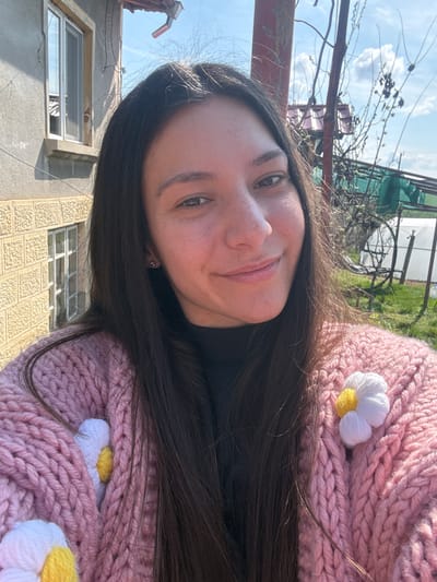 A woman is posing for a selfie outdoors. She has long, dark hair and fair skin with freckles. She is wearing a chunky knit sweater in a pale pink color, adorned with crocheted daisies.

The background includes a building with light-colored walls and barred windows, suggesting a residential area. A metal fence and branches of trees are also visible. The presence of a greenhouse or covered structure, in addition to the building style, implies that this image was taken in a rural location within the city of Lipnik, Bulgaria. The lighting suggests it is daytime.