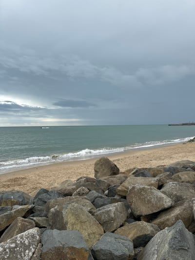 The image shows a beach scene in Courtown, Ireland, under a cloudy sky. In the foreground, large, grey and brown rocks are piled along the shoreline. A sandy beach stretches out to the sea, where gentle waves are breaking. The sea is a murky green color, and a distant pier or jetty can be seen on the right horizon. A small object, possibly a jet ski, is visible in the middle distance on the water. The overall atmosphere is overcast and somber.