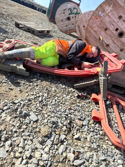 In Ballyboughal, Ireland, an outdoor scene on a gravel and dirt surface depicts a male individual, appearing to be asleep or resting, lying prone on a red plastic construction barrier. He is dressed in fluorescent yellow work trousers, an orange high-visibility vest over a dark long-sleeved shirt, and muddy work boots. A smartphone is visible in his right hand. The environment includes additional red plastic barriers, two wooden pallets, and three large wooden cable spools, two marked "General Cable" with one numbered "22," positioned in the background. A green and red container is partially visible behind the spools. The scene is illuminated by direct sunlight under a clear sky.